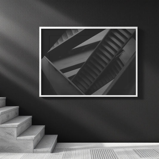 A framed black and white architectural photograph of a spiral staircase, displayed on a dark wall above a staircase in a modern interior setting.