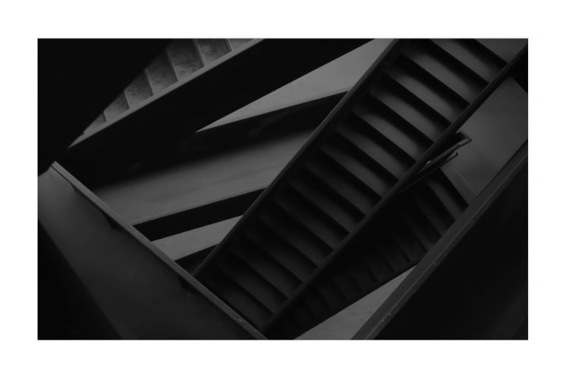 Black and white architectural photograph of a modern staircase with strong diagonal lines and layered steps, presented as a square art print with a white border.