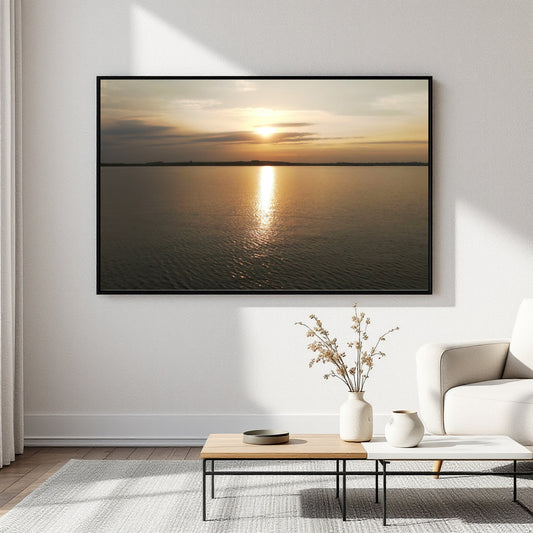 A framed horizontal canvas print of a sunset over calm water, displayed on a white wall in a modern living room setting above a coffee table and sofa.