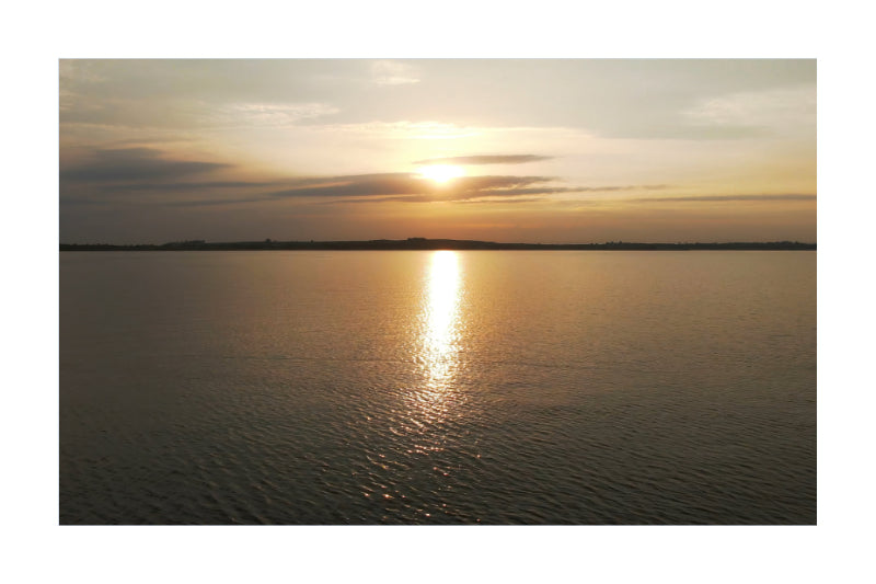 A scenic sunset over a calm body of water, with the sun low on the horizon, golden light reflecting vertically across the water surface, and a dark shoreline in the distance.