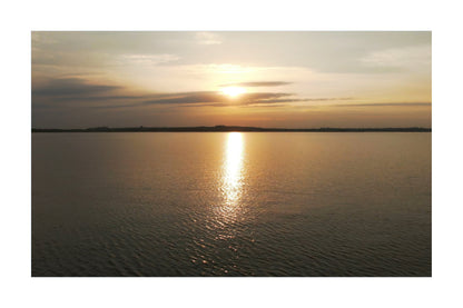 A scenic sunset over a calm body of water, with the sun low on the horizon, golden light reflecting vertically across the water surface, and a dark shoreline in the distance.