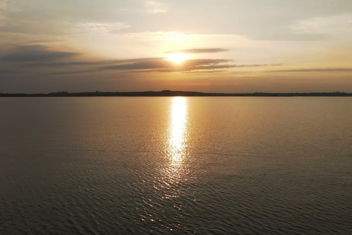A scenic sunset over a calm body of water, with the sun low on the horizon, a bright reflection of golden light on the water, and soft clouds in the sky.