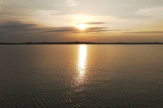 A scenic sunset over a calm body of water, with the sun low on the horizon, a bright reflection of golden light on the water, and soft clouds in the sky.
