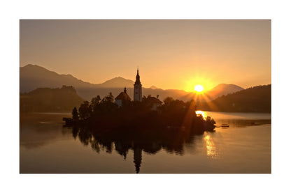 A scenic landscape print of an island with a church spire on a lake at sunset, with mountains in the background and the sun reflecting on the water.
