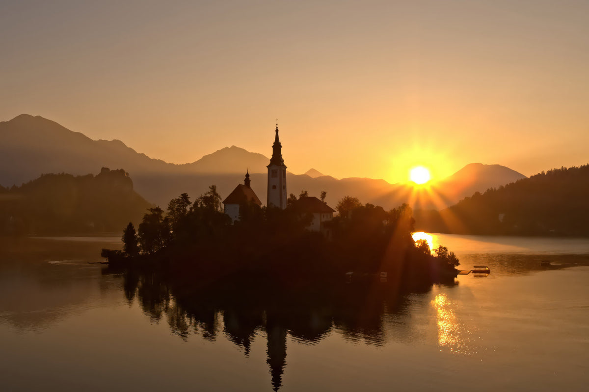 A scenic landscape photograph of Bled Lake in Slovenia at sunset, featuring the silhouette of the Church of Our Lady of Mount Carmel on an island, with mountains in the background and the sun reflecting on the water.