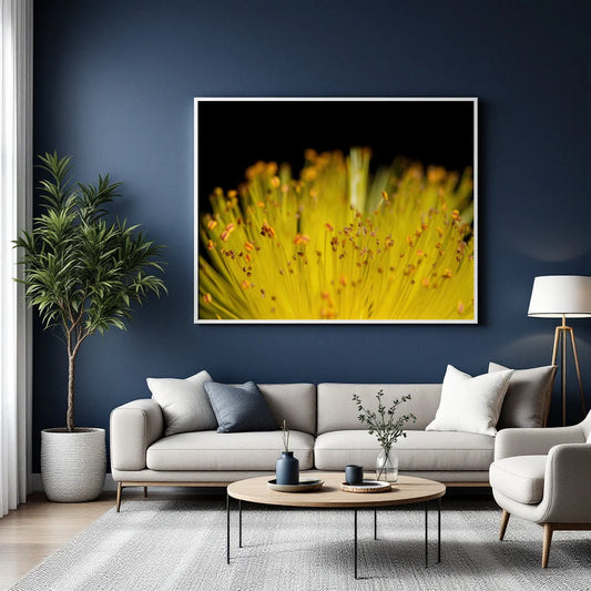 A framed wall art print featuring a close-up yellow flower on a black background, displayed above a light gray sofa in a modern living room with a blue wall, potted plant, coffee table, and floor lamp.