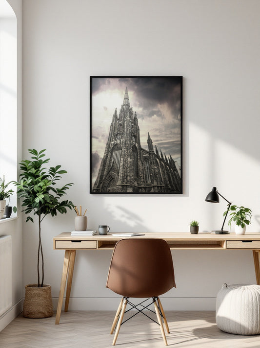 A framed black and white art print of a Gothic cathedral spire hangs on a white wall above a wooden desk in a modern, sunlit room with a brown chair and plants.