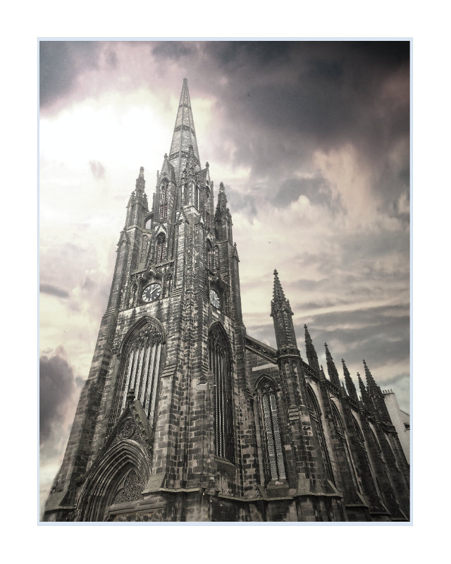 A framed art print depicting a dramatic, low-angle view of a Gothic cathedral spire against a cloudy, overcast sky, presented with a white border.