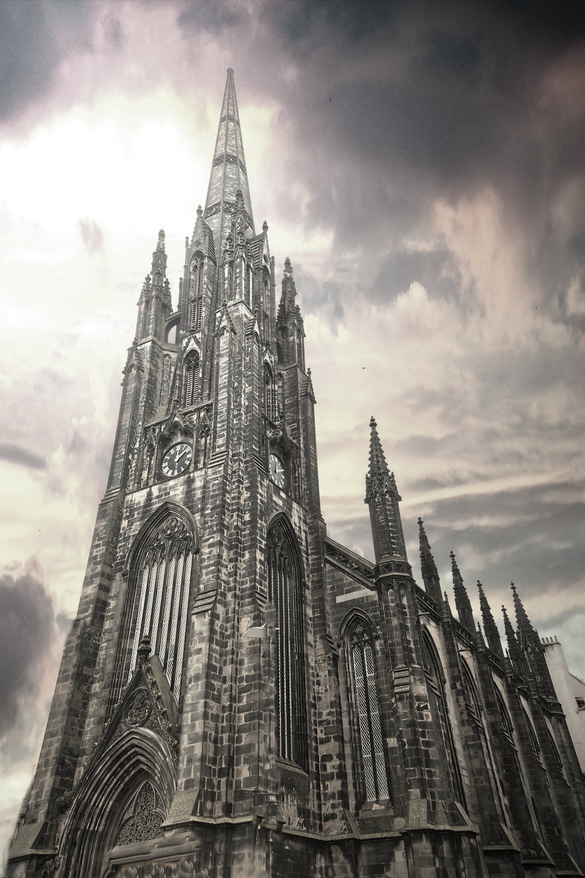 A dramatic, low-angle photograph of a Gothic cathedral spire against a cloudy, overcast sky, rendered in a high-contrast black and white or sepia-toned monochrome style.