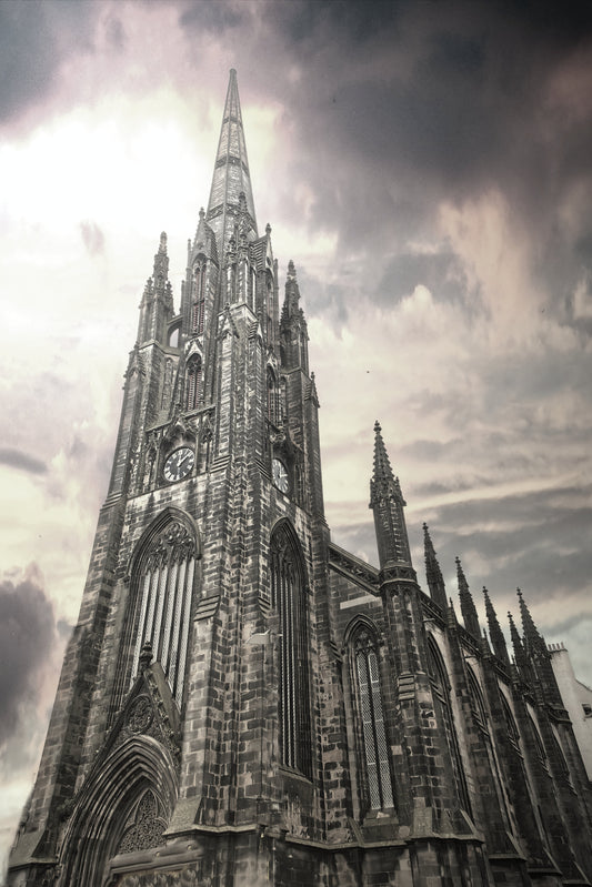 A dramatic, low-angle photograph of a Gothic cathedral spire against a cloudy, overcast sky, rendered in a high-contrast black and white or sepia-toned monochrome style.