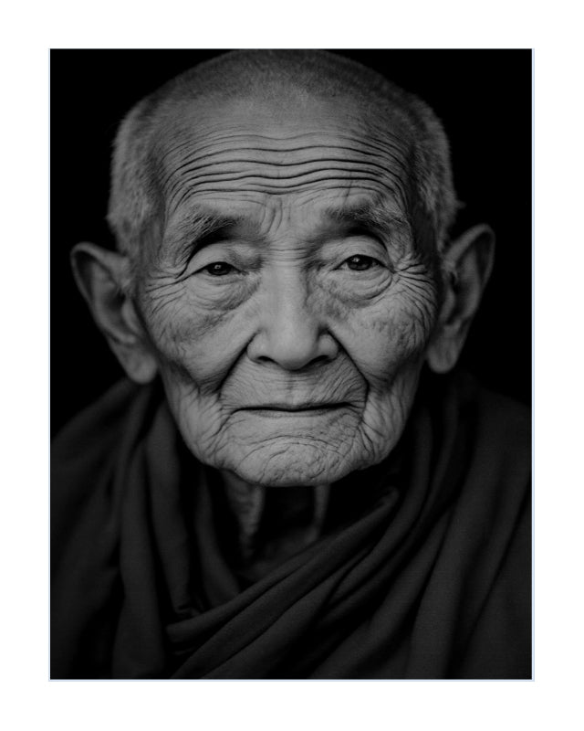 Black and white portrait print of an elderly buddhist woman with deep facial wrinkles, wearing a dark draped garment against a black background.