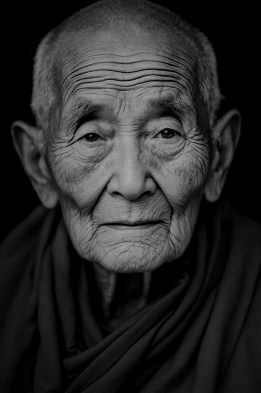 Black and white close-up portrait of an elderly buddhist woman with deeply wrinkled skin, wearing a dark draped garment against a solid black background.