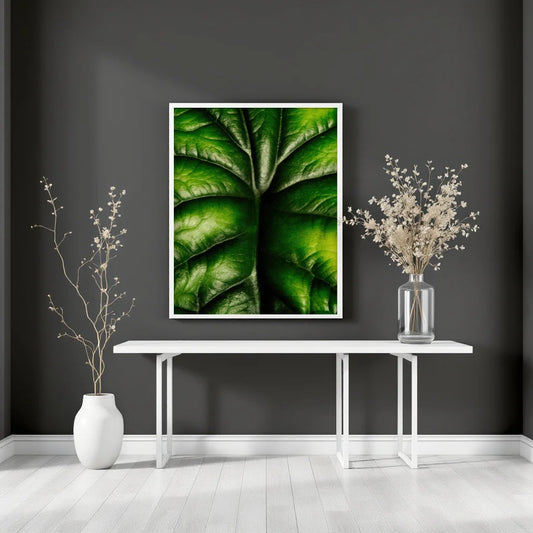 A framed botanical art print of a green leaf hangs on a dark gray wall above a white console table, styled with two white vases containing dried branches in a bright, modern room.
