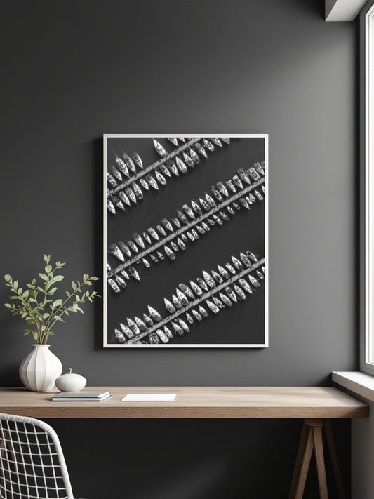 A framed black and white botanical art print featuring diagonal rows of feather-like shapes, displayed on a dark gray wall above a wooden desk with a vase and plant.