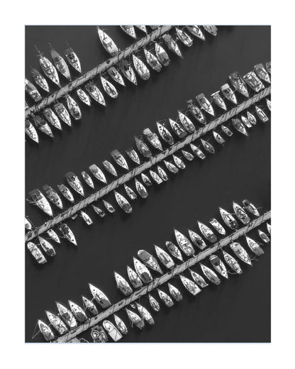 Black and white photographic print depicting multiple rows of small boats tied side-by-side along diagonal ropes on a dark water surface.