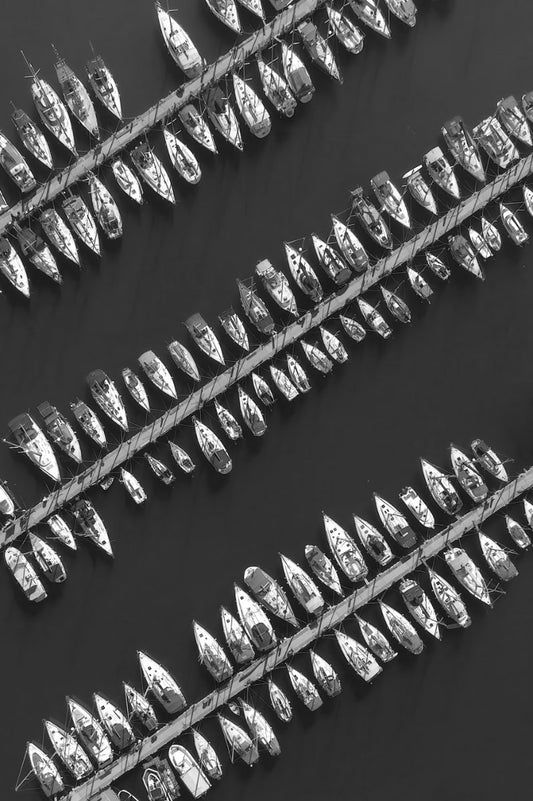 Black and white aerial-style photograph of many small boats tied side by side in long diagonal rows on a dark water surface.