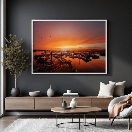 A large framed art print depicting a vibrant orange and pink sunset over a marina, displayed on a dark charcoal wall above a modern wooden console table in a styled living room.