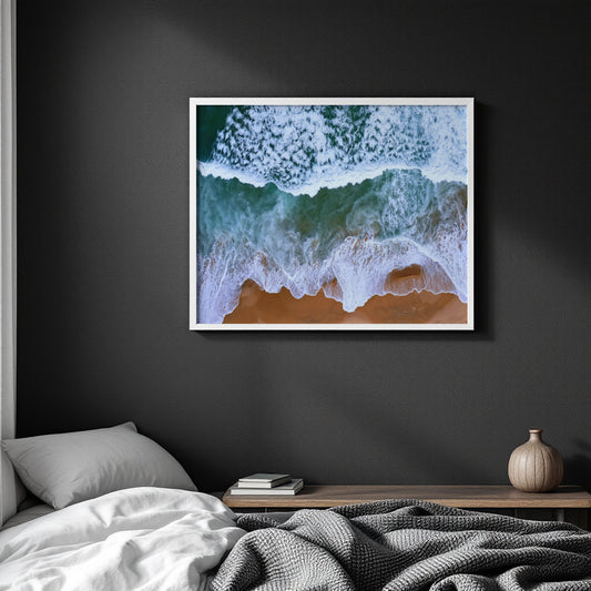 A framed square wall art print showing ocean waves breaking on a sandy beach, displayed above a bed in a dark modern bedroom.