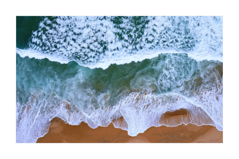 Aerial view of ocean waves with white foam crashing onto a sandy beach, shown as a square print centered on a white background.