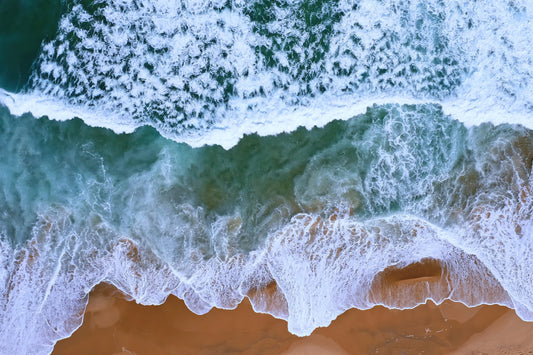 Aerial view of ocean waves with white foam crashing onto a sandy beach, showing turquoise and deep blue-green water.