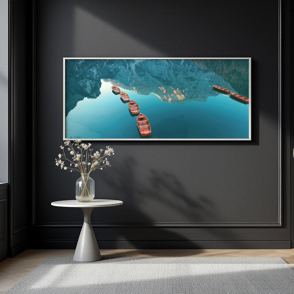 A framed horizontal wall art print showing an aerial view of blue water with red boats, displayed on a dark charcoal wall above a small white side table with a vase of dried flowers.