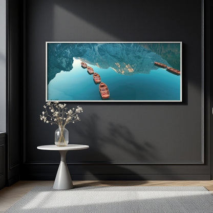 A framed horizontal wall art print showing an aerial view of blue water with red boats, displayed on a dark charcoal wall above a small white side table with a vase of dried flowers.