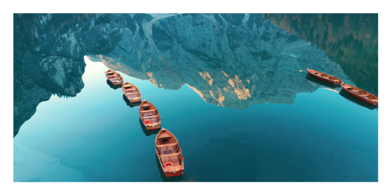 A scenic photograph of several small wooden boats floating on calm blue water, with a large mountain range reflected in the lake.