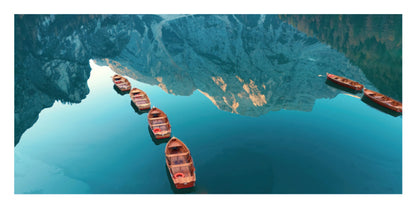 A scenic photograph of several small wooden boats floating on calm blue water, with a large mountain range reflected in the lake.