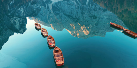 A scenic aerial view of a calm blue lake reflecting jagged mountains, with several small reddish-brown boats floating on the water.