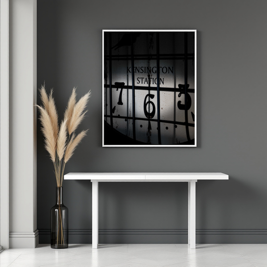 A framed black-and-white art print of a vintage train station clock face with the text 'KINGSINGTON STATION' hangs on a dark gray wall above a white console table, with a vase of pampas grass to the left.