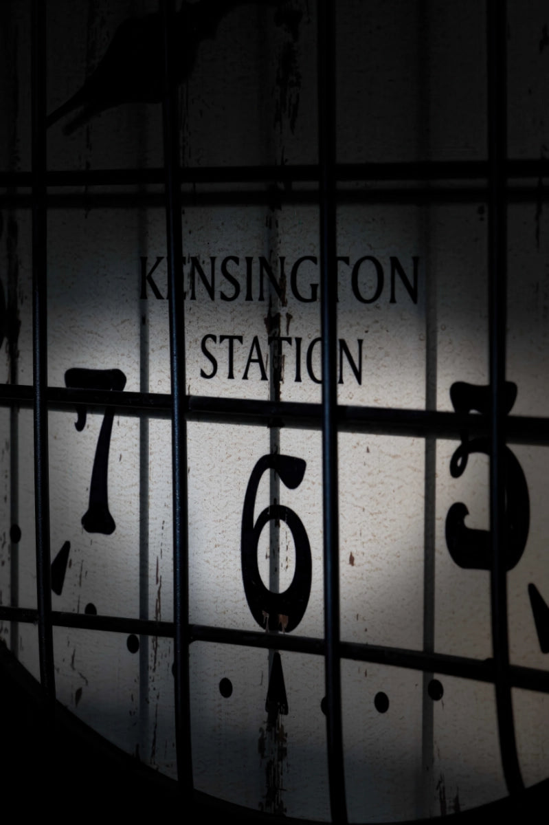 A dark, moody graphic featuring the words 'KENSINGTON STATION' in black serif text over a gray background, layered behind a black grid pattern with clock-style numbers 7, 6, and 5.