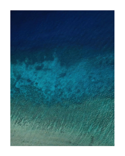 Three distinct bands of blue run across clear ocean water in an aerial view with a white mount border around.