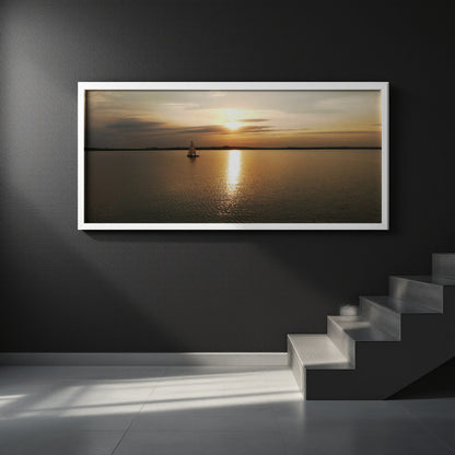 A framed horizontal landscape print of a sunset over water with a small sailboat, displayed on a dark gray wall above a staircase.