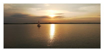 A scenic sunset over calm water with a small sailboat near the center and the sun low on the horizon, shown in a wide landscape frame with a white border.