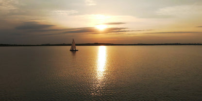 A scenic sunset over a calm lake with a small sailboat silhouette on the water and the sun low on the horizon, casting a bright golden reflection across the surface.