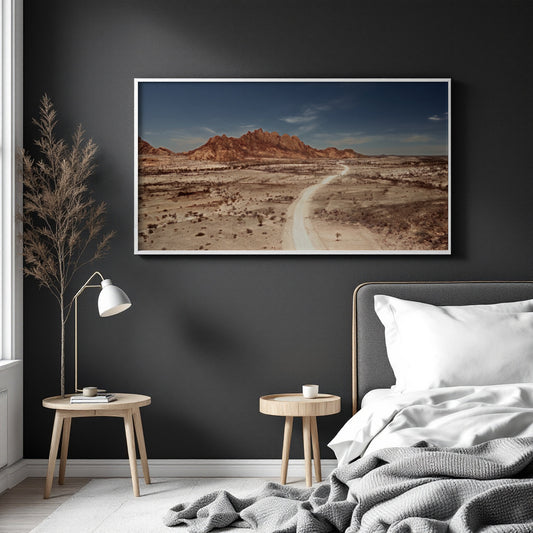 A framed landscape art print of a desert canyon with a winding dirt road, displayed on a dark charcoal wall above a bed in a modern bedroom setting.