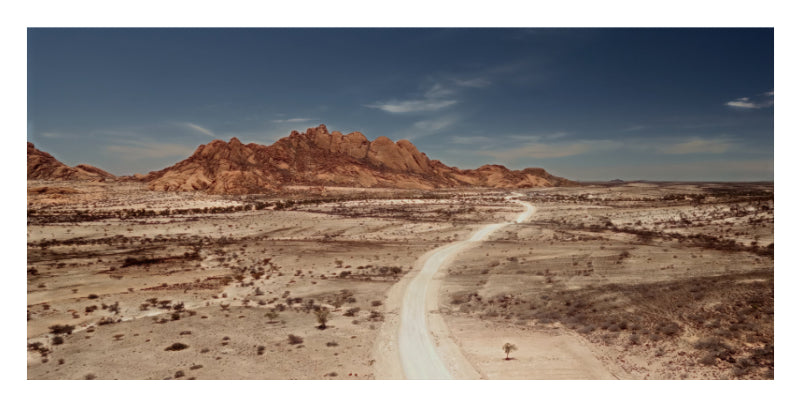 A wide desert landscape with a winding pale road through sandy terrain and a prominent reddish rock formation under a deep blue sky.