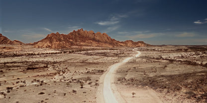Aerial view of a desert landscape with a winding dirt road leading toward rugged red rock mountains under a deep blue sky.
