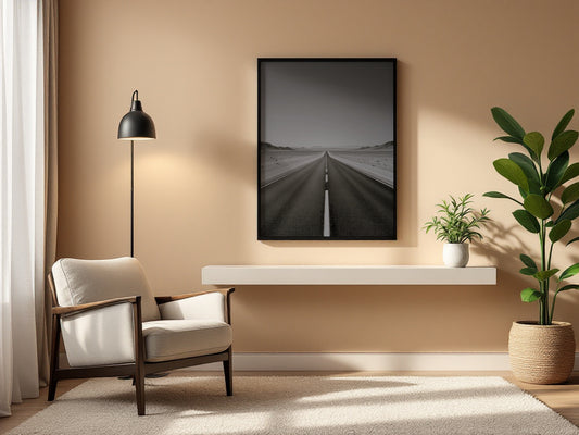 A framed black and white landscape photograph of a straight road through a desert hangs on a beige wall in a modern living room setting with a white armchair, floor lamp, and potted plant.