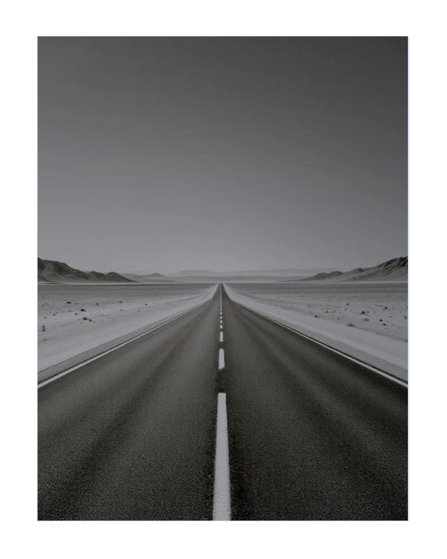 A black and white landscape photograph of a straight paved road leading toward distant mountains, shown as a vertical art print with a white border.