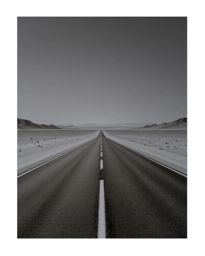 A black and white landscape photograph of a straight paved road leading toward distant mountains, shown as a vertical art print with a white border.