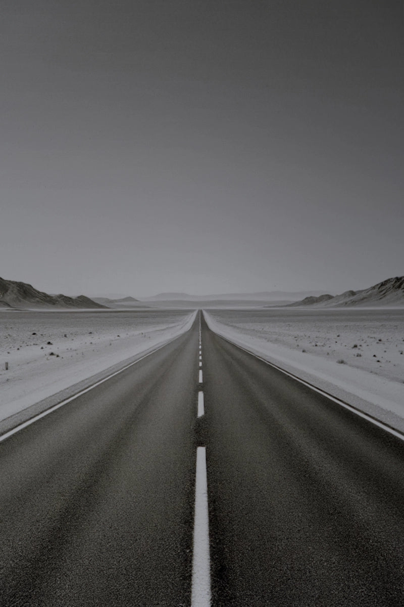 A straight paved road runs through a flat desert landscape under a pale gray sky, with low hills visible on both sides and a dashed white line down the center.