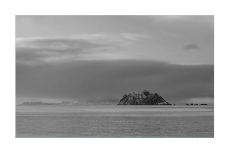 Black and white landscape photograph of a small rocky island in calm water under a cloudy sky, presented as a square print with a white border.
