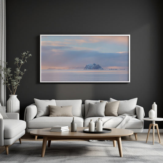 A framed horizontal landscape art print depicting a misty mountain island under a soft pastel sky, displayed on a dark charcoal wall above a modern light gray sofa in a styled living room.