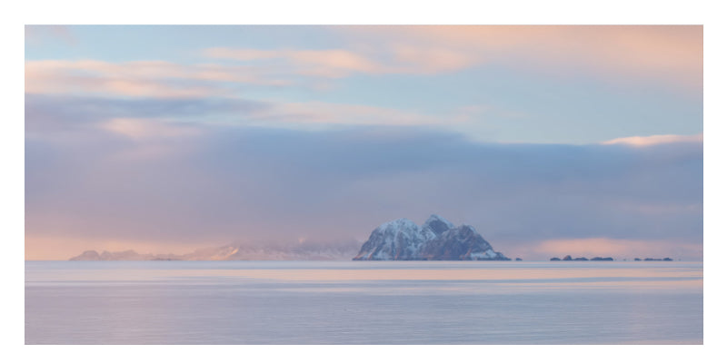 A scenic landscape photograph of a small snow-capped island or rock formation rising from calm water under a soft pastel sky with pink, blue, and lavender tones.