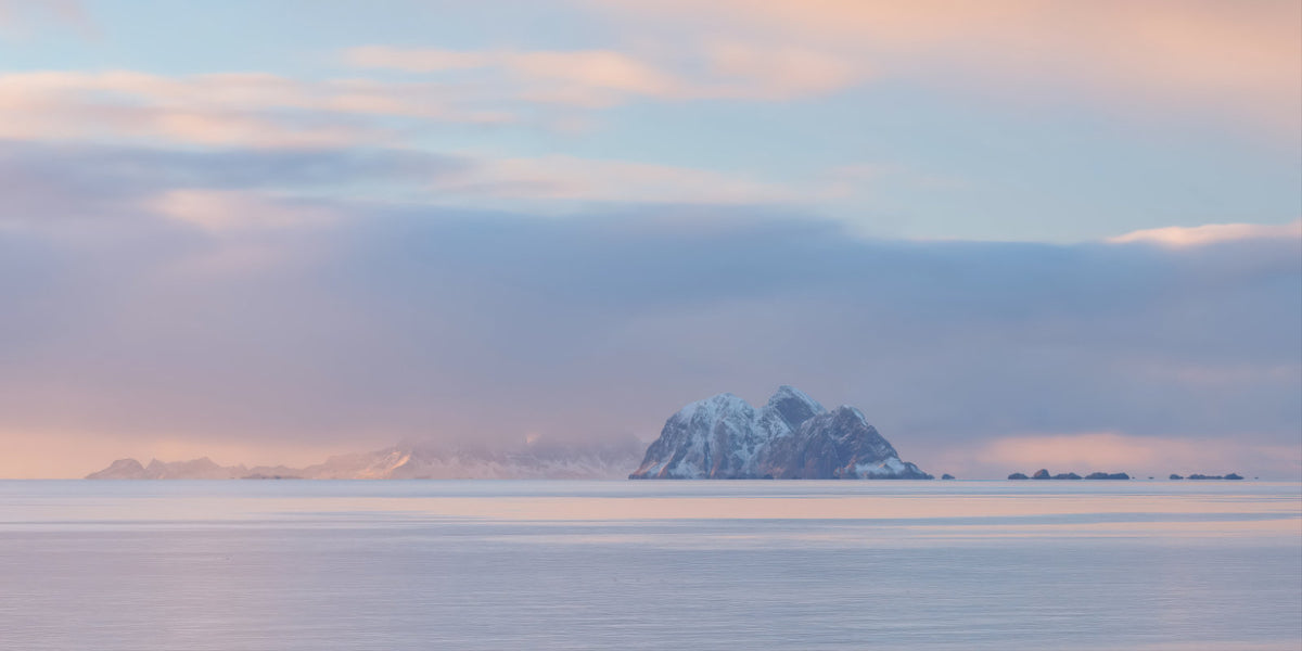 A serene landscape photograph of a snow-capped mountain island in a calm sea under a soft pastel sky with pink, blue, and gray tones.