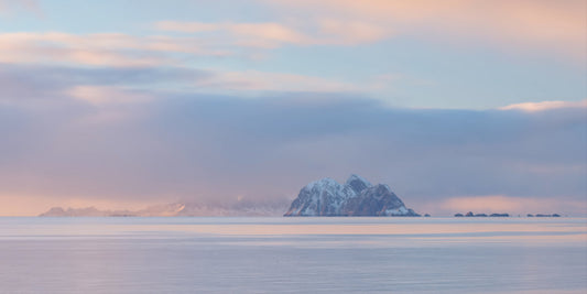 A serene landscape photograph of a snow-capped mountain island in a calm sea under a soft pastel sky with pink, blue, and gray tones.