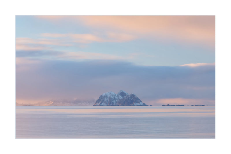 A serene landscape photograph of a snow-capped mountain island in a calm body of water under a soft pastel sky with pink, peach, and blue tones.