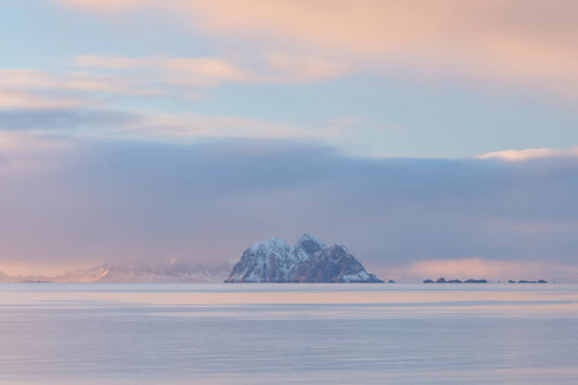A calm seascape with a small snow-capped island or rock formation in the center distance, under a pale blue sky with soft pink and peach clouds.