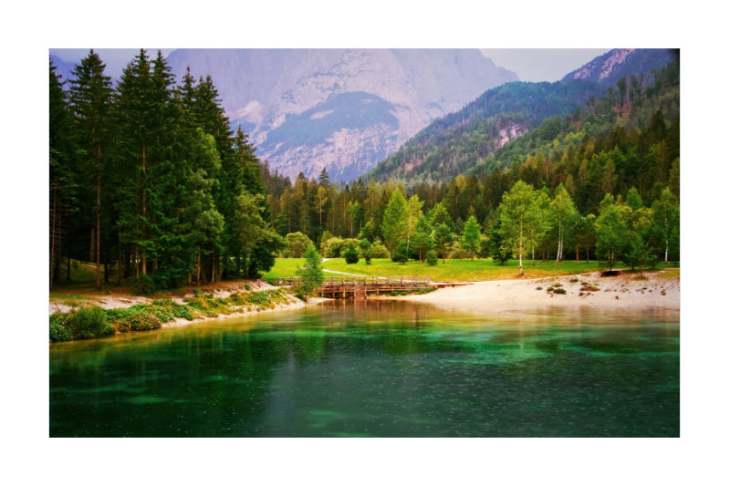 A scenic landscape print featuring a clear green lake in the foreground, a sandy shore lined with trees, a grassy meadow, and tall evergreen forests leading up to rugged mountains in the background.